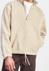 Fleecejacka i beige med halvmidja, texturerat tyg, ribbade ärmslut och dragsnodd i nederkanten. Sidopaneler i en ljusare nyans.