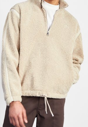 Veste en polaire beige avec un zip sur la moitié du devant, tissu texturé, poignets côtelés et ourlet avec cordon de serrage. Panneaux latéraux dans une teinte plus claire.