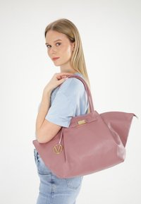 Rosa Leder-Tasche mit strukturiertem Finish, ausgestattet mit einem goldfarbenen Hardwareverschluss und einem kleinen dekorativen Anhänger, über der Schulter getragen.