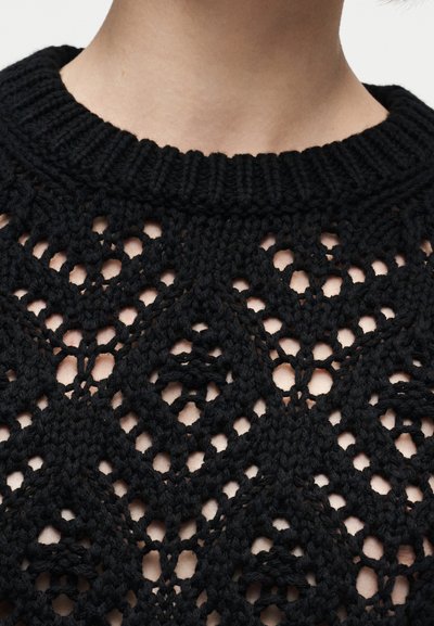 Prim-plan al unei persoane care poartă un pulover negru tricotat, cu guler nervurat și un model deschis țesut în formă de diamant.