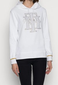 Felpa bianca con cappuccio e logo in strass, caratterizzata da un tocco d'oro sui polsini e una vestibilità rilassata. Tessuto morbido e liscio con corde regolabili.