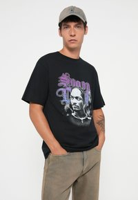 Sort bomulds t-shirt med korte ærmer, der har et grafisk print af en mand med fletninger og tekst i lilla. Båret med lysebrune bukser.