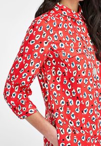 Chemise rouge avec un motif léopard, tissu léger, deux poches poitrine et un col avec manches longues retroussées.