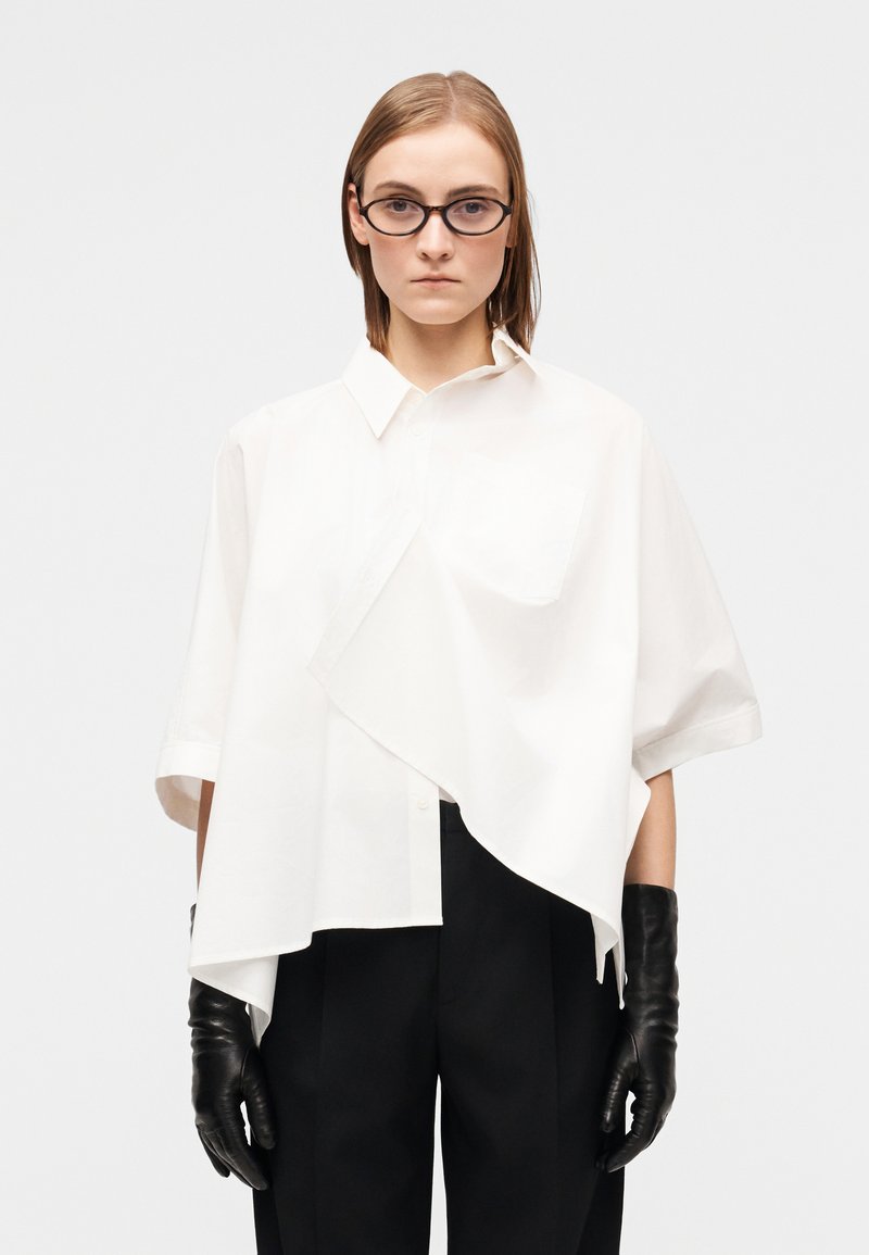 Chemise blanche oversize à boutons avec ourlet asymétrique et poche, associée à des gants en cuir noir et un pantalon noir ajusté.