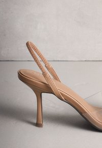 Chaussure slingback beige en cuir à talon haut avec panneau latéral transparent et talon aiguille fin sur une surface unie.