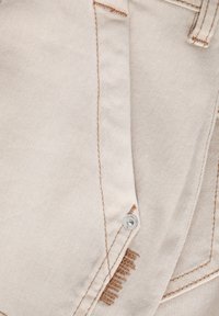 Primer plano de una tela de mezclilla beige con costuras marrones visibles, un remache plateado y parte de un detalle del bolsillo trasero.