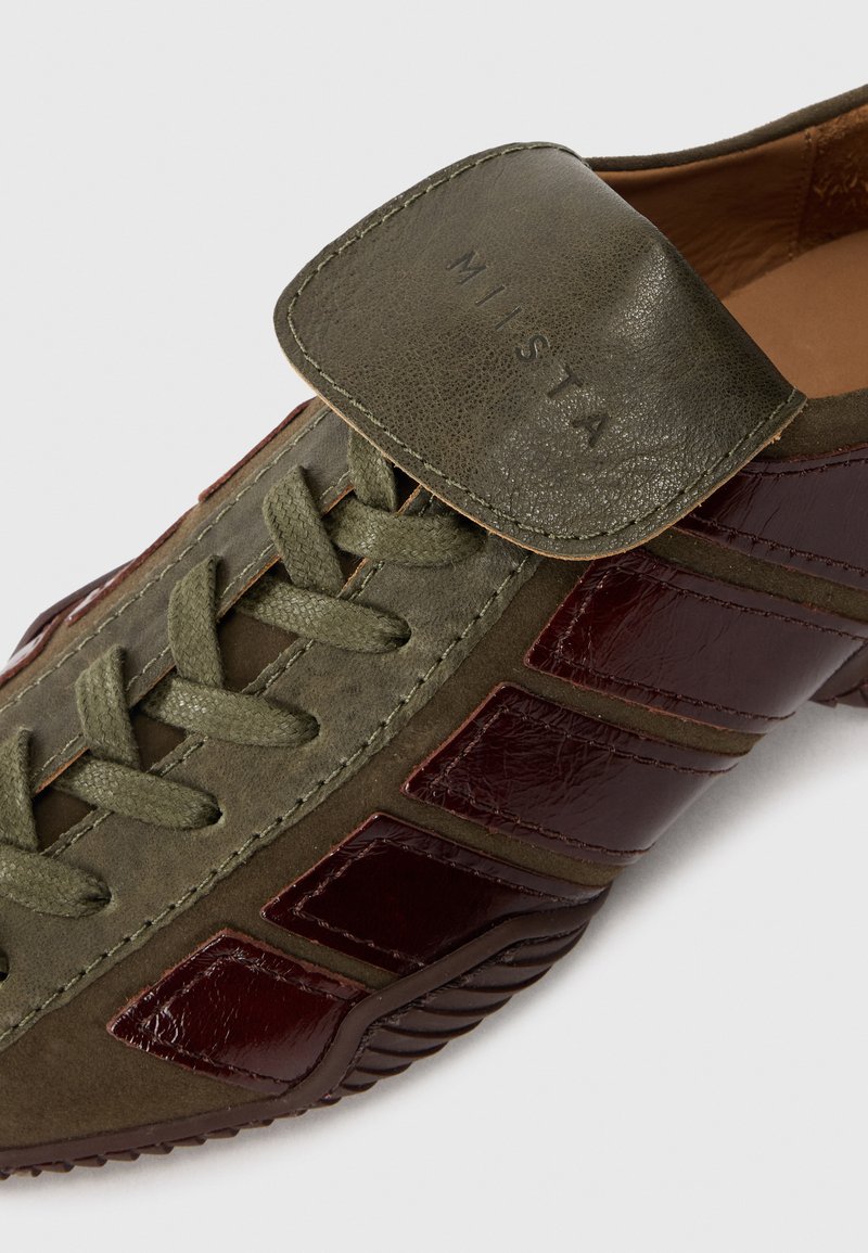 Green leather sneaker with burgundy patent accents, featuring a thick sole, lace-up design, and a flap with embossed brand logo.