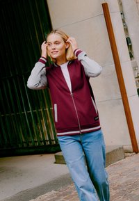 Un sweat à capuche zippé bordeaux avec des manches grises, des rayures blanches aux poignets et des poches à l'avant. Porté par-dessus une chemise blanche et associé à un jean bleu clair.