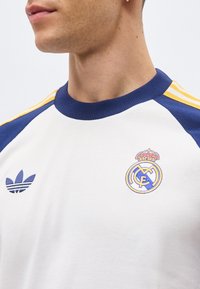 Homme portant une chemise blanche et bleue à rayures jaunes, avec le logo Adidas à gauche et le blason du Real Madrid sur la poitrine droite.