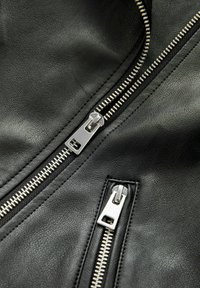 Detailní pohled na dva stříbrné zipy na texturované černé kožené tkanině, jeden větší a jeden menší, uspořádané diagonálně.