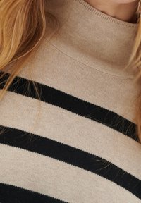 Nahaufnahme eines beigen und schwarzen gestreiften Rollkragenpullovers, getragen von einer Person mit langen blonden Haaren.