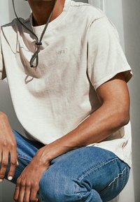 Beżowa koszulka z krótkim rękawem z małym logo Levi's, w połączeniu z niebieskimi dżinsami o średnim odcieniu. Charakteryzuje się luźnym krojem i miękką teksturą.