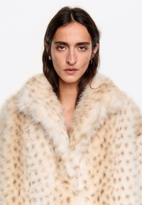 Abrigo de piel sintética en crema con patrón de manchas en marrón claro, con un cuello ancho y una superficie suave y texturizada. Vista frontal centrada.