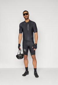 Schwarzes Radsport-Outfit mit kurzen Ärmeln, figurbetontem Design und Reißverschluss vorne. Schwarzer Helm in der Hand, Sporthandschuhe, schwarze Schuhe und Logo-Akzente.