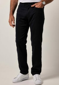 Pantaloni in cotone blu navy con design aderente, dotati di tasche frontali e chiusura con zip. Indossati con sneakers bianche e un orologio d'argento.