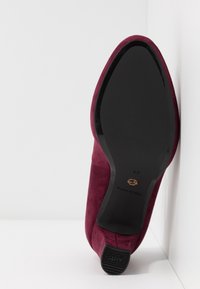 Chaussure pour femme en daim marron vue en angle, semelle vers le haut, montrant une semelle noire lisse et un embout de talon en caoutchouc « antidérapant », taille 37, sur fond blanc.