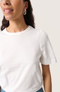 Witte T-shirt met korte mouwen en een ronde halslijn, licht gepofte mouwen, gemaakt van zachte katoenstof, aangevuld met zilveren oorbellen in de vorm van een ring.