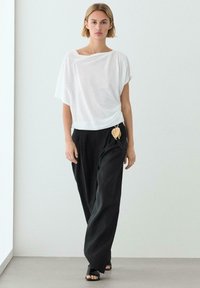 Donna che cammina al chiuso indossando una maglia bianca ampia, pantaloni neri larghi con un accessorio a forma di foglia e sandali neri aperti sulle dita.