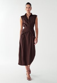 Robe marron sans manches avec un col boutonné, une ceinture à la taille et deux grandes poches à l'avant. Le tissu semble texturé et tombe jusqu'au mollet.