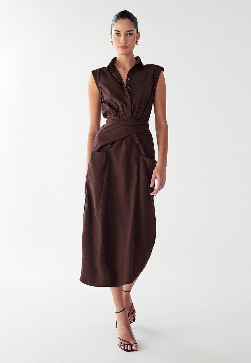Robe marron sans manches avec un col boutonné, une ceinture à la taille et deux grandes poches à l'avant. Le tissu semble texturé et tombe jusqu'au mollet.
