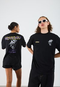Black cotton t-shirts feature graphic designs in white and purple. One shirt has a floral motif, while the other displays stylized text.