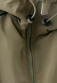 Zbliżenie na zamek kurtki w oliwkowym kolorze z czarnymi zaciągaczami i metką z napisem "Bershka" na kapturze.