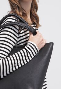 Svart läder tote-väska med sydda handtag, slät textur och en överdimensionerad form. Modellen bär en svart och vit randig långärmad skjorta.