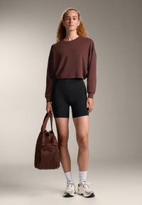 Brauner, gekürzter Sweatshirt, schwarze Radshorts und eine braune Tasche. Weiße Socken und mehrfarbige Sneakers runden den Look ab. Glatte Texturen überall.