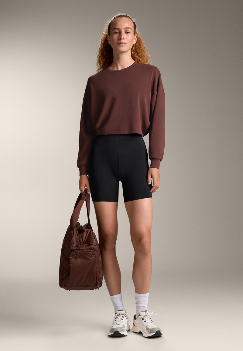 Brauner, gekürzter Sweatshirt, schwarze Radshorts und eine braune Tasche. Weiße Socken und mehrfarbige Sneakers runden den Look ab. Glatte Texturen überall.