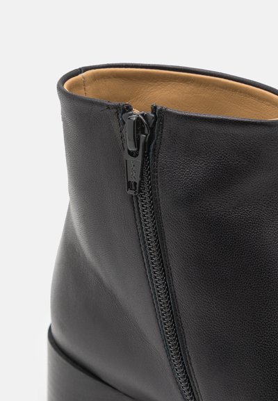 Bota de tornozelo em couro preto com fecho lateral, textura suave, forma arredondada e um salto baixo empilhado. Interior forrado com material em tom claro.