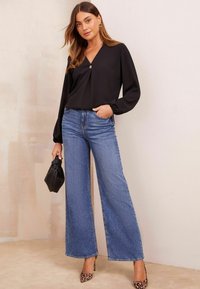 Maglietta nera a maniche lunghe con scollo a V e un bottone dorato, abbinata a jeans in denim blu a gamba larga e una borsa nera testurizzata.