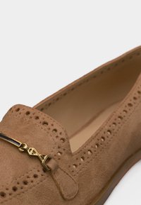Mocassin en daim marron avec perforations décoratives et ornement en métal doré à l'avant supérieur.