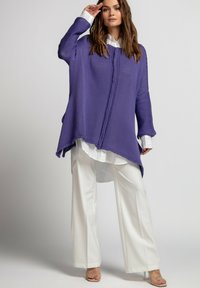 Fioletowy, oversize'owy sweter tunikowy z teksturowanym wzorem, z długimi rękawami i asymetrycznym dolnym brzegiem, noszony na białej koszuli z guzikami i szerokich spodniach.