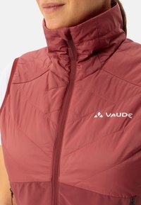 Gilet isolato rosso con colletto alto, motivo trapuntato, zip frontale e logo "VAUDE" bianco sul petto sinistro. Texture liscia e lucida.