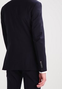 Marinblå kostymjacka med en enkel design, enkel slits bak och detaljer med fyra knappar vid ärmslutet. Tillverkad av ett slätt tyg med minimal textur och rena linjer.