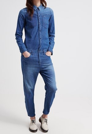 Kobieta ubrana w niebieski dżinsowy kombinezon z długimi rękawami, z przodu zapięty na guziki, w białe buty sznurowane, stojąca z rękami w kieszeniach.