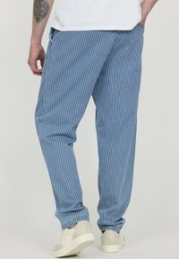 Un pantalon rayé bleu en tissu léger, présentant des rayures blanches verticales et une coupe décontractée, associé à des baskets blanches.