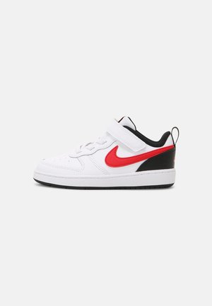 Biały dziecięcy sneaker z czerwonym logo Nike, czarną piętą i paskiem na rzep na jednolitym jasnoszarym tle.