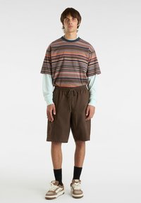 Wzorzysty T-shirt z krótkim rękawem w różnych kolorach noszony na jasnoniebieskiej koszuli z długim rękawem, w połączeniu z brązowymi szortami do kolan i trampkami.