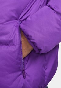 Giacca a vento viola con superficie testurizzata, dotata di una tasca con zip e polsini aderenti. Il materiale sembra leggero e isolante.