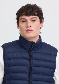 Tânăr cu păr scurt și închis la culoare, purtând o geacă matlasată fără mâneci, bleumarin, peste o cămașă albă, pe un fundal deschis.