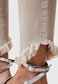 Pantalones vaqueros beige con dobladillos deshilachados, combinados con pulseras metálicas plateadas en los tobillos. Tela texturizada con un ligero brillo en el dobladillo.