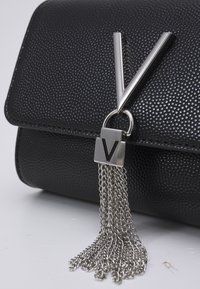 Sac à main en cuir noir texturé avec un logo 'V' métallique et un détail de pompon en chaîne. La fermeture à rabat offre un design épuré.