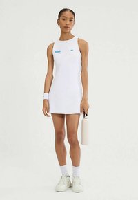 Weißes ärmelloses Kleid mit blauen Logoakzenten. Glatter Stoff, figurbetontes Design und gerader Schnitt. Hält eine hellfarbige Wasserflasche.