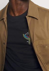 Camicia beige con bottoni sopra un maglione nero. Il maglione presenta ricami di uccelli colorati e un collettino tondo.