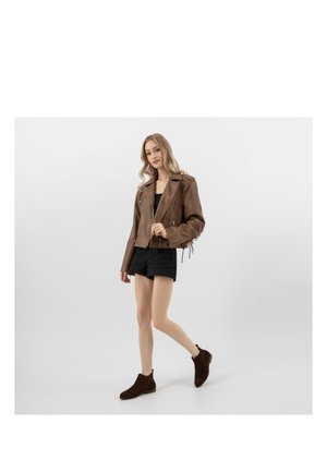 Žena kráčející, oblečená v hnědé kožené bundě, černých šortkách, černém topu a tmavě hnědých kotníkových botách na bílém pozadí.