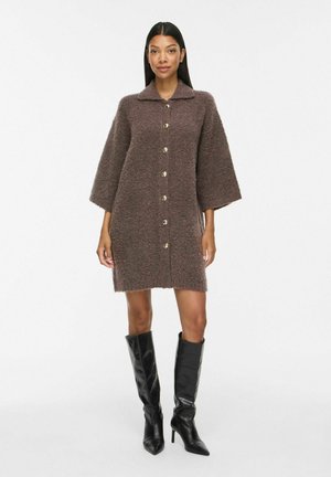 Femme portant une robe marron texturée à manches longues larges avec boutons et des bottes noires à talons montant jusqu'aux genoux, debout sur un fond blanc.