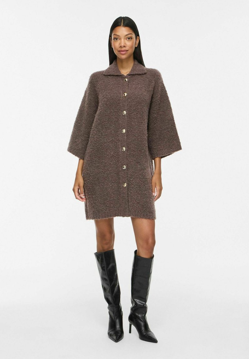 Femme portant une robe marron texturée à manches longues larges avec boutons et des bottes noires à talons montant jusqu'aux genoux, debout sur un fond blanc.