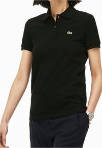 Polo noir en coton avec un col, des manches courtes et un logo Lacoste vert sur la poitrine gauche. Porté avec un pantalon foncé.