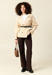 Beige dubbelknäppt trench coat med bälte, bärs över en vit topp. Bruna mönstrade utsvängda byxor och svarta kraftiga stövlar. Brun handväska.